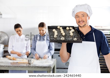 Portrait of smiling chef carrying baking sheet with dough balls while female colleagues working in background at commercial kitchen