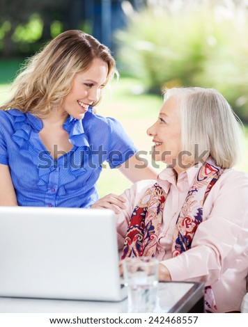 Happy granddaughter and grandmother looking at each other while using laptop on nursing home porch