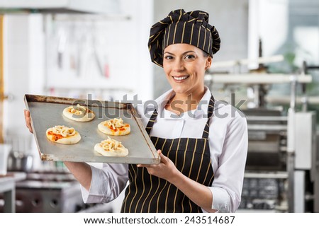 Portrait of happy female chef holding small pizzas on baking sheet in commercial kitchen