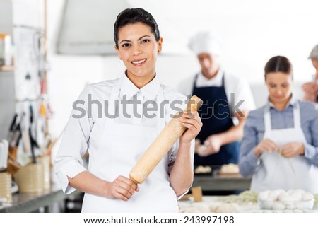 Portrait of smiling female chef holding rolling pin while colleagues preparing pasta at commercial kitchen