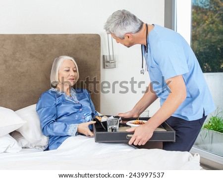 Male nurse serving breakfast to senior woman on bed in nursing home