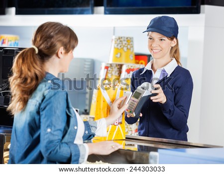 Smiling female worker accepting payment from woman through NFC technology at cinema concession stand