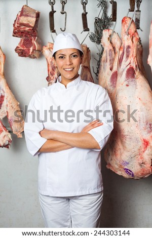 Portrait of confident female butcher standing against meat hanging in butchery
