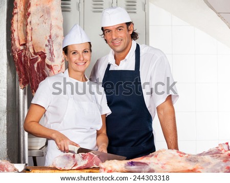 Portrait of confident female butcher cutting meat while standing by male colleague at counter in shop