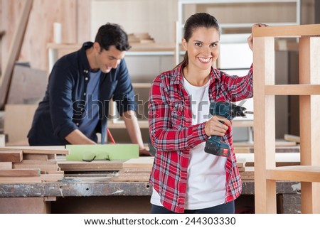 Portrait of happy female carpenter drilling wood while colleague working in background at workshop