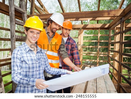 Portrait of confident female architect with male colleagues examining blueprint in wooden cabin at site