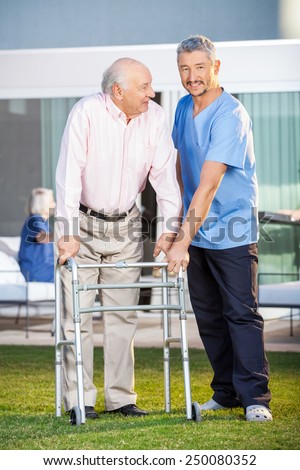 Portrait of smiling caretaker assisting senior man to use walking frame at nursing home lawn