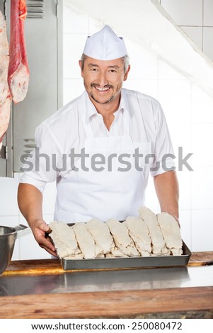 Portrait of confident mature butcher holding tray filled with dredge chicken in shop