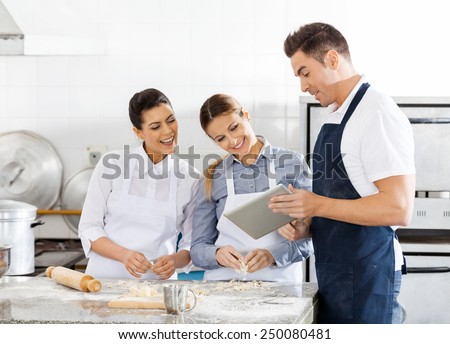 Happy chefs checking recipe on digital tablet while preparing pasta in commercial kitchen