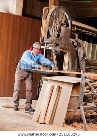 Full length portrait of happy senior male carpenter using bandsaw in workshop