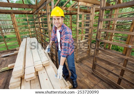 Portrait of smiling carpenter measuring wooden plank with tape in cabin at construction site