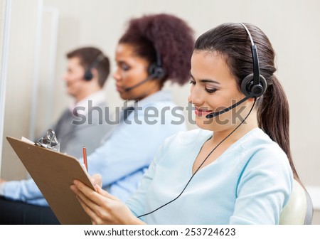Smiling female customer service representative writing on clipboard with colleagues in background at office