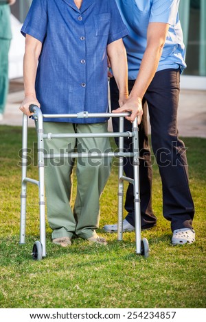 Low section of male caretaker helping senior woman in using walking frame at nursing home lawn