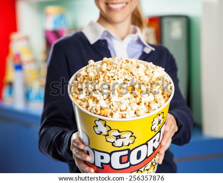 Midsection of smiling female worker offering popcorn bucket at cinema concession stand
