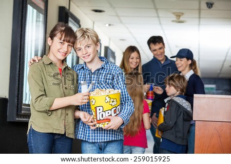 Portrait of happy brother and sister holding popcorn at cinema with family and worker in background