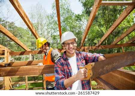 Portrait of happy construction worker cutting wood with handsaw while colleague in background at site