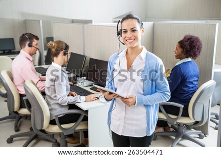 Portrait of happy female customer service representative holding tablet computer while colleagues working in background at call center