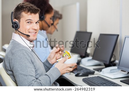 Portrait of young smiling male customer service representative having food while colleagues working in office