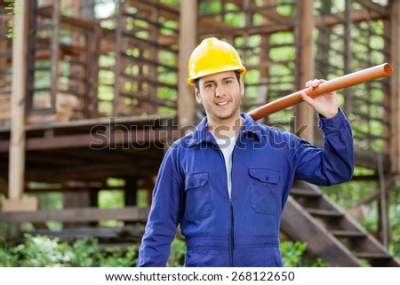 Portrait of smiling male worker holding pipe against incomplete timber cabin at construction site