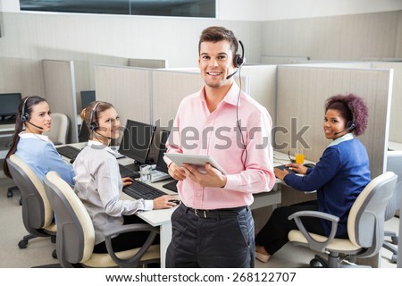Portrait of young male customer service executive holding tablet computer while colleagues looking at him in call center