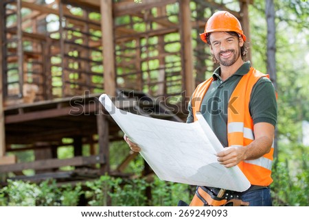 Portrait of happy male architect holding blueprint against incomplete timber cabin at construction site