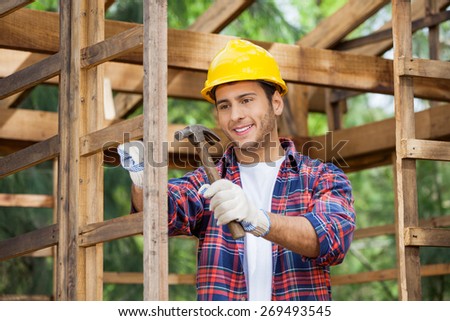 Smiling male worker hammering nail on wooden cabin at construction site