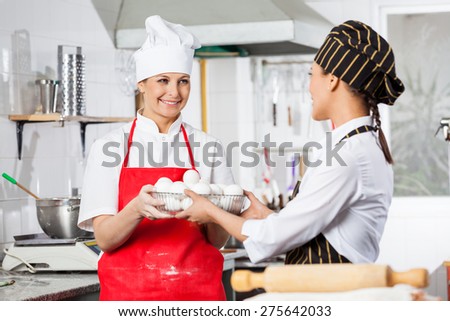 Happy female chef giving container full of eggs to colleague in commercial kitchen