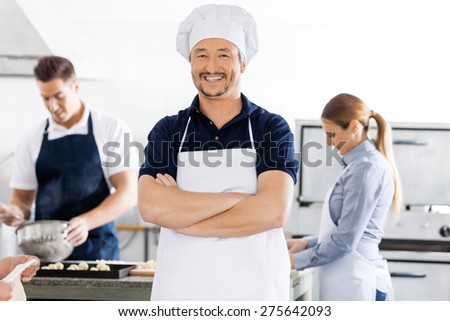 Portrait of confident male chef standing arms crossed while colleagues working in background at commercial kitchen