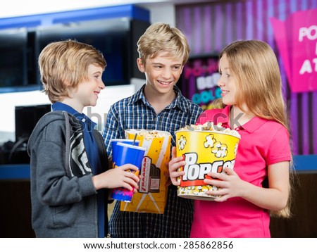 Happy brothers and sister conversing while holding snacks against cinema concession stand