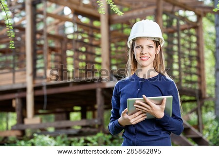 Portrait of smiling female architect holding digital tablet against incomplete timber cabin at construction site