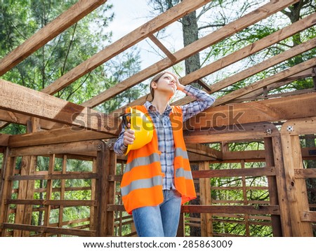 Tired female construction worker holding hardhat while standing in incomplete timber cabin at site