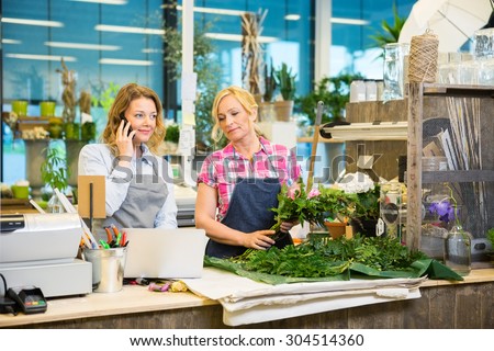 Florist using mobile phone while colleague making bouquet at counter in flower shop