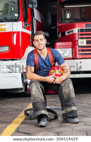 Full length portrait of smiling firefighter holding helmet while sitting against trucks at fire station