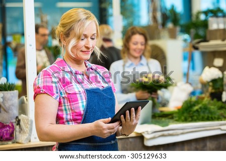 Smiling mature female florist using digital tablet with colleague working in background at shop