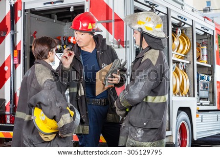 Male firefighter discussing with colleagues while holding clipboard against truck at fire station