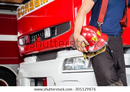 Midsection of young fireman holding red helmet while leaning on firetruck at station