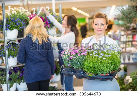 Portrait of female florist carrying flower plants with colleague assisting customer in background at shop