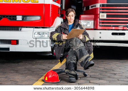 Full length young firewoman writing on clipboard while sitting against trucks at fire station