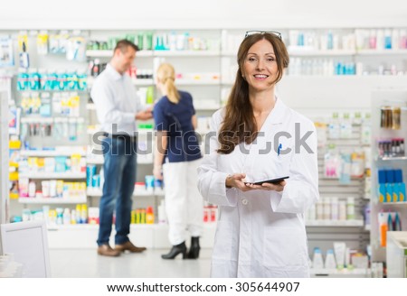 Portrait of smiling female chemist holding digital tablet while assistant and customer standing in background at pharmacy