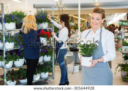 Portrait of happy florist holding flower pot with colleague assisting customer in background at shop