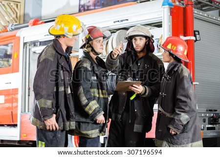 Young male firefighter showing something to colleagues against truck at fire station