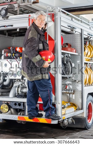 Full length portrait of smiling fireman standing on truck at fire station