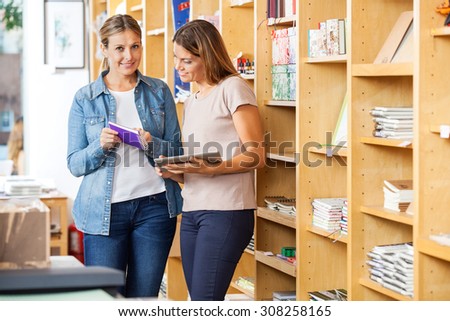 Portrait of smiling customer holding book while checking list with friend on digital tablet at store