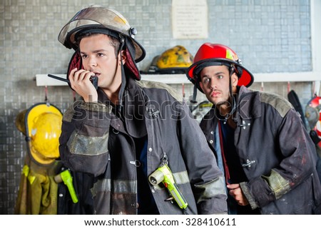 Male firefighter using walkie talkie at fire station with colleague standing in background