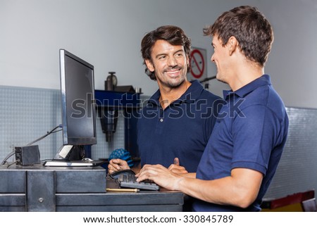 Happy male mechanic discussing with colleague while using computer in garage