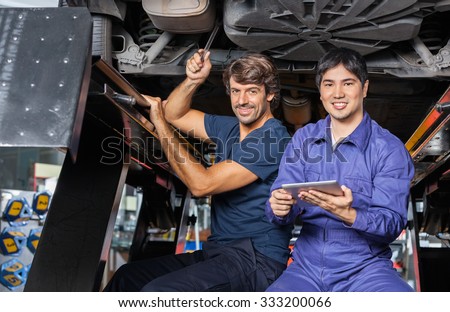 Portrait of smiling mechanics with digital tablet working under lifted car at auto repair shop