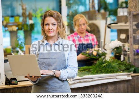 Portrait of female florist using laptop with colleague working in background at flower shop