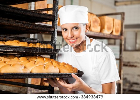 Female Baker Holding Baking Tray By Rack In Bakery