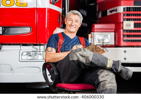 Smiling Firefighter Sitting On Chair Against Trucks