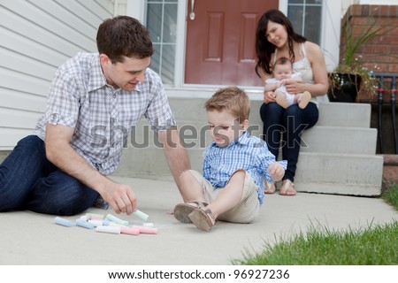 Young Father And Son Drawing With Chalk And Mother Sitting With Baby At Background In Front Of Their House.